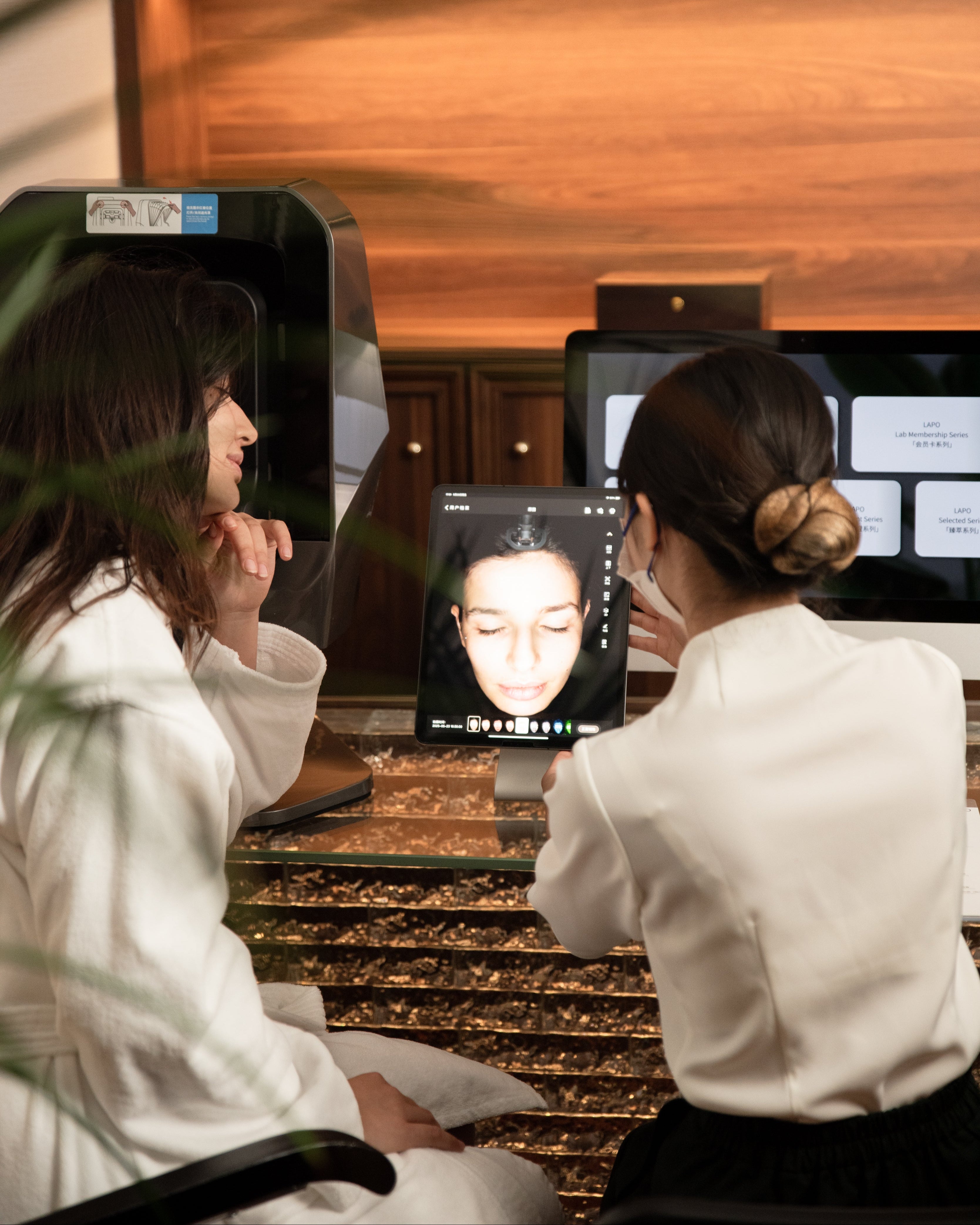 Person receiving a facial treatment with a technician using a computer screen displaying facial analysis software.