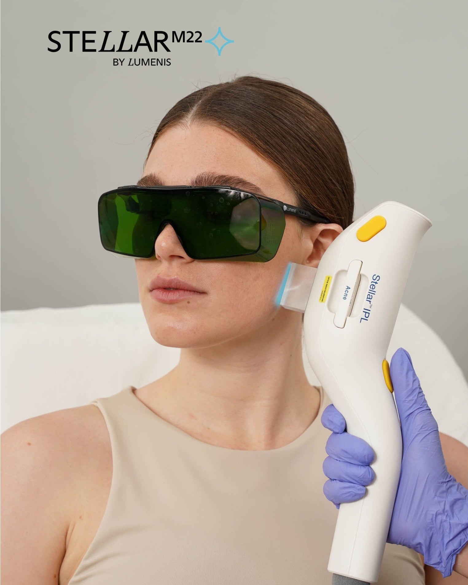 Person wearing green laser safety glasses with a Stellar M22 device held by a gloved hand against a gray background.